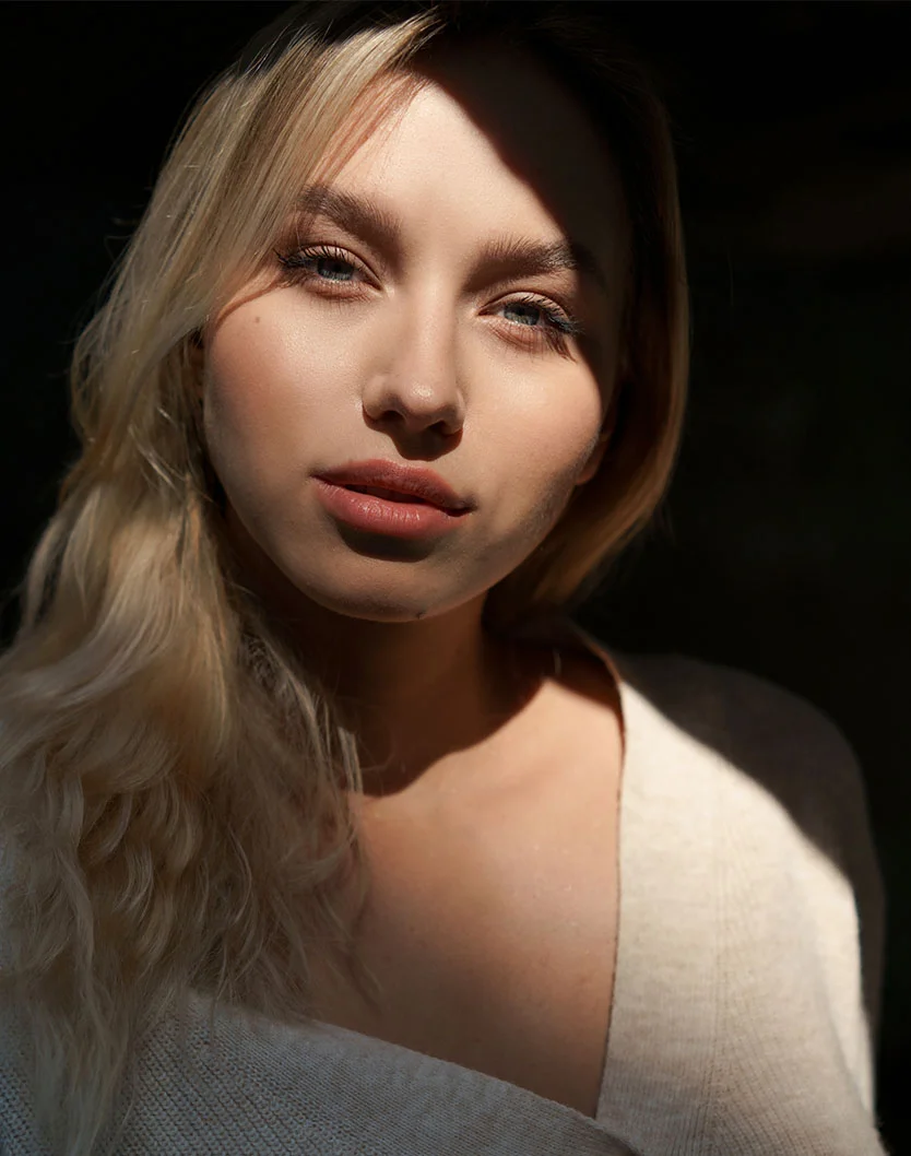 A portrait of a young woman with long blonde hair and blue eyes, looking directly at the camera. Sunlight illuminates her face from the right, creating a strong contrast of light and shadow. She is wearing a light-colored, knitted sweater. - BBL Hero in Los Angeles, CA