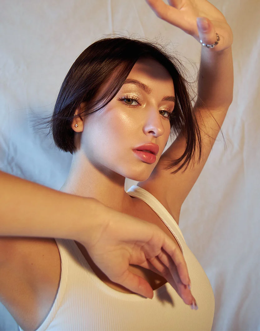 A medium-shot portrait of a young woman with short, dark hair and shimmery gold eyeshadow. She is wearing a white tank top and has both arms raised with her hands near her face, as if dancing. She is looking directly at the camera with a neutral expression. - Bellafil Bellalift in Los Angeles, CA