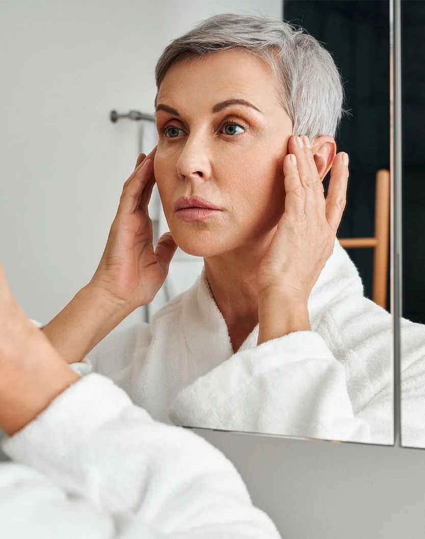 A woman with short, gray hair is looking at her reflection in a mirror. She is wearing a white bathrobe. She is touching her face, and her expression appears somewhat concerned or pensive. The lighting in the bathroom is bright and neutral. The background is a light gray/white bathroom wall. The reflection in the mirror shows a similar-aged woman with a similar expression. - Collagen Stimulation in Los Angeles, CA