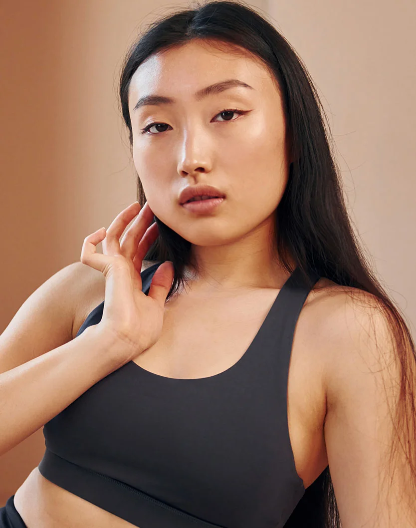 A head-and-shoulders portrait of a young woman with long, dark hair, wearing a dark gray sports bra. She is looking directly at the camera with a neutral expression and has her left hand gently touching her face. - Daxxify in Los Angeles, CA