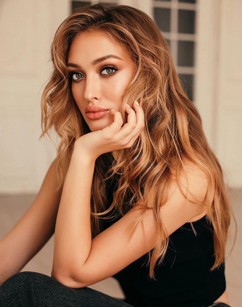 A head-and-shoulders portrait of a young woman with long, wavy blonde hair and blue eyes. She is wearing a black top and is resting her chin on her hand, looking directly at the camera. She has full lips and a neutral expression. - Dermal Fillers including all brands RHA in Los Angeles, CA