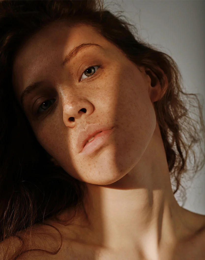 Close-up of a woman with shoulder-length, wavy dark brown hair and light tan skin with freckles across her nose and cheeks. Soft lighting casts shadows that highlight her features. She has light green eyes and natural rosy pink lips. The background is softly blurred, emphasizing her face and neck in a natural, gentle style. - Dermaplaning in Los Angeles, CA