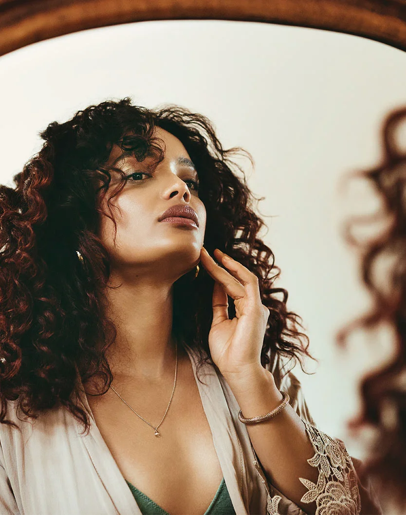 A young woman with long, curly brown hair, wearing a beige robe with lace trim. She is looking at her reflection in a mirror and touching her chin with her right hand. She is wearing a thin gold necklace and a beaded bracelet. - Facials in Los Angeles, CA