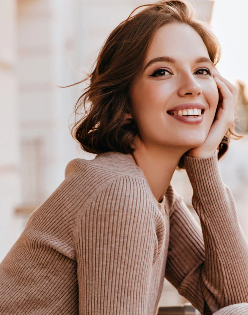 A smiling young woman with short brown hair. She is wearing a beige ribbed sweater and resting her chin on her hand, looking off to the side. The background is a soft, out-of-focus outdoor setting. - Glacial RX in Los Angeles, CA