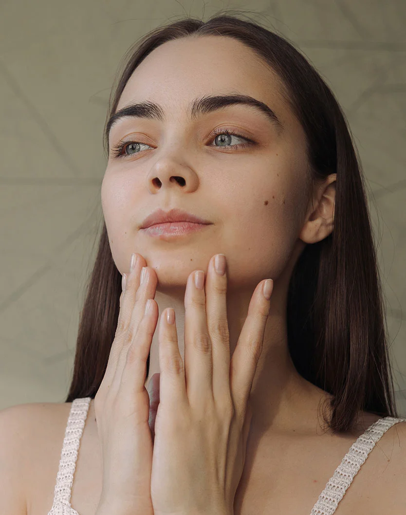 A close-up portrait of a young woman with long dark hair and blue eyes, gently touching her face with both hands. She is wearing a light-colored, lacy top and looking to the left, away from the camera. Her skin appears smooth and luminous. - Halo Laser in Los Angeles, CA