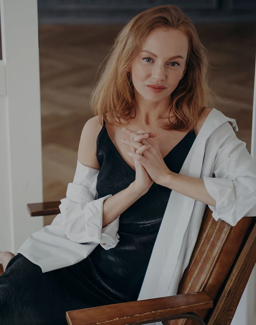 A woman with light reddish-brown hair and freckles is sitting in a wooden chair. She is wearing a black slip dress with a white button-down shirt draped over her shoulders. She has her hands clasped together in her lap and is looking directly at the camera with a slight smile. - Hand Rejuvenation in Los Angeles, CA