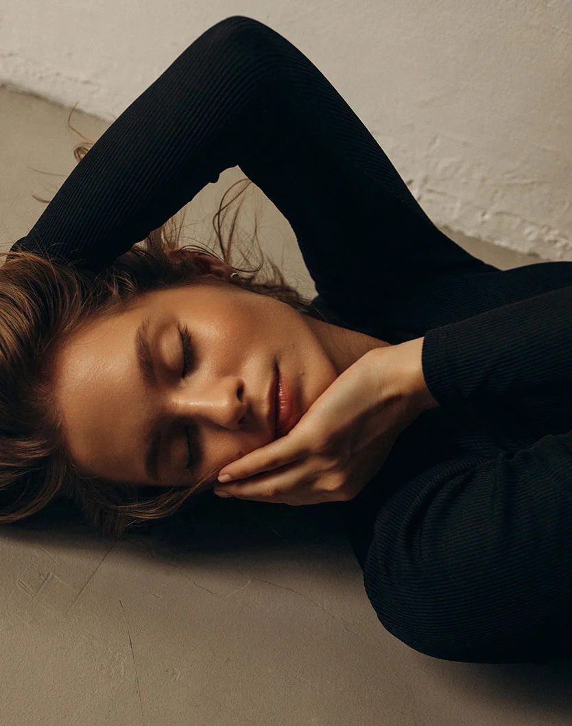A young woman with light brown hair, wearing a black long-sleeved shirt, lying on her side on a gray surface with her head resting on her arms and looking directly at the camera, eyes closed. - Hydrafacial in Los Angeles, CA
