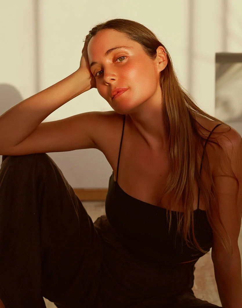 A woman with long light brown hair sits, resting her head on her right hand. She wears a black sleeveless top and dark pants, with sun-kissed skin and a calm, pensive expression. Warm, natural light casts gentle shadows on her and the light beige background. - Laser Skin Rejuvenation in Los Angeles, CA