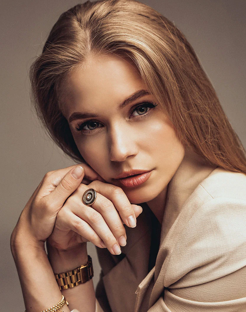 A young woman with long, blonde hair, wearing a beige blazer. She is seated, looking directly at the camera with her hands clasped together near her chin. She is wearing a gold watch, two gold bracelets, and a ring with a large black and gold design. The background is a solid, light gray. - Lip Flip Removal in Los Angeles, CA