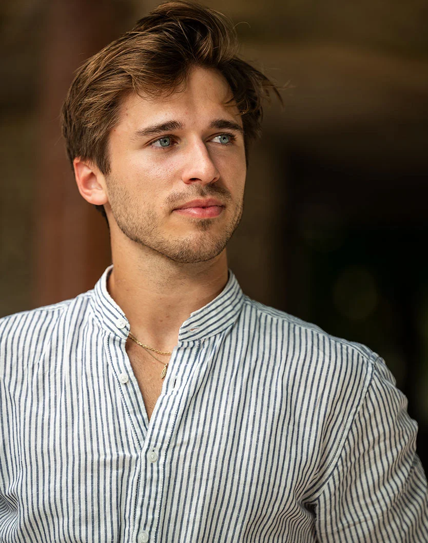 Close-up of a young man with light brown, slightly tousled hair and light blue eyes. He has a short, trimmed beard and a light complexion. He wears a light-colored, striped button-down shirt with a mandarin collar and a gold necklace. He looks off to his right against a warm, softly blurred background. - Male Penile Enhancement in Los Angeles, CA