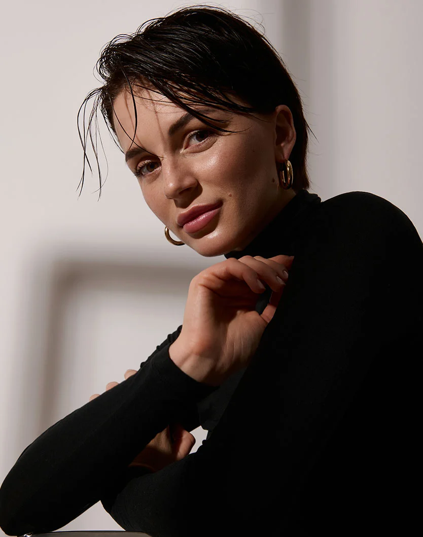 Close-up of a woman with short, slightly damp dark hair. She has light tan skin and wears a black turtleneck and gold hoop earrings. Her expression is neutral with a hint of a smile. Hands are partially visible on her torso. The background is off-white with soft shadows. - PDO Threads in Los Angeles, CA