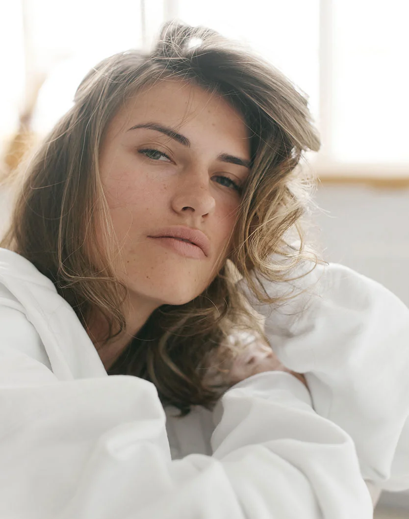 Close-up of a young woman with light brown, slightly tousled hair framing her face. Her expression is neutral and contemplative, with her gaze directed slightly off to the viewer’s right. She has smooth, light-toned skin and wears a white, loose-fitting top. Soft, diffused lighting creates a gentle glow around her, with a window or light source visible in the bright, airy background.- Peels in Los Angeles, CA