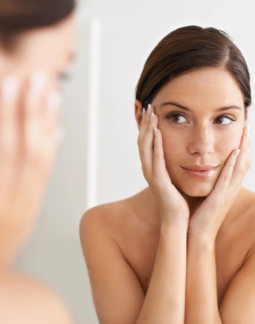 A young, light-skinned woman with shoulder-length brown hair looks at herself in a mirror, gently touching her cheeks with both hands. Her expression is thoughtful or pleased. The mirror reflection is softly blurred, and the light beige background with soft, natural lighting suggests a bathroom-like setting. - PRP in Los Angeles, CA