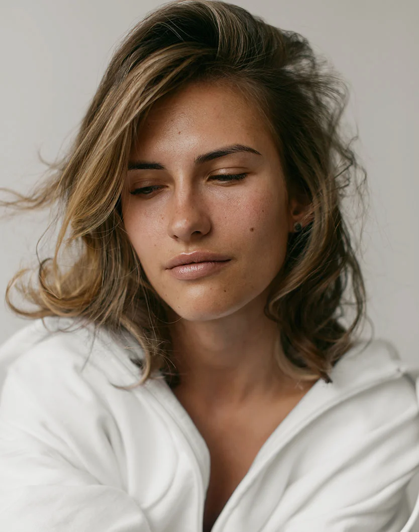 A close-up of a light-skinned woman with shoulder-length, slightly tousled light brown hair. She looks downward with a contemplative expression. Her skin is smooth with a light tan tone and visible freckles, and her lips are a soft neutral pink. She wears a loose, white, textured garment draped around her shoulders. The light gray background and soft, diffused lighting create a gentle, understated mood. - Radiesse in Los Angeles, CA