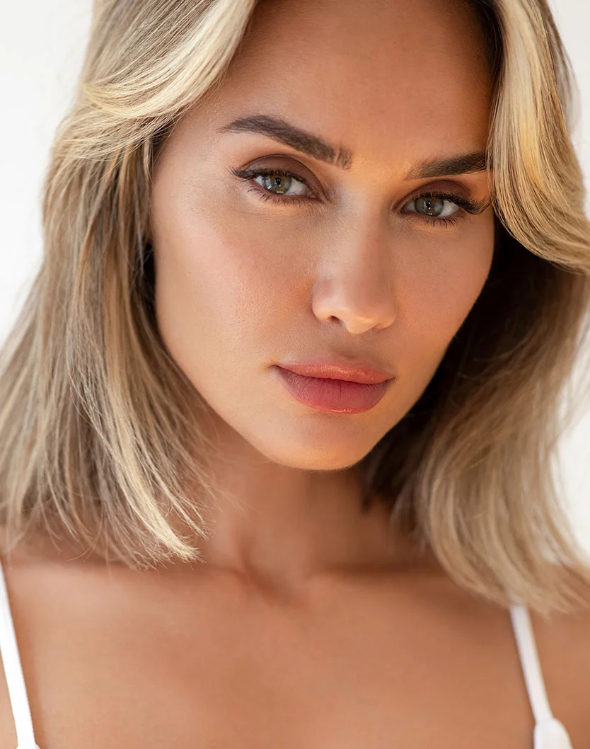 A close-up of a woman with light blonde, slightly wavy hair and a neutral, direct gaze. Her skin is smooth and light-toned, with defined eyebrows and pinkish-nude lipstick. She wears a white sleeveless top or bra. Soft lighting highlights her eyes and lips, focusing attention on her face and upper body. - Red YAG Laser in Los Angeles, CA