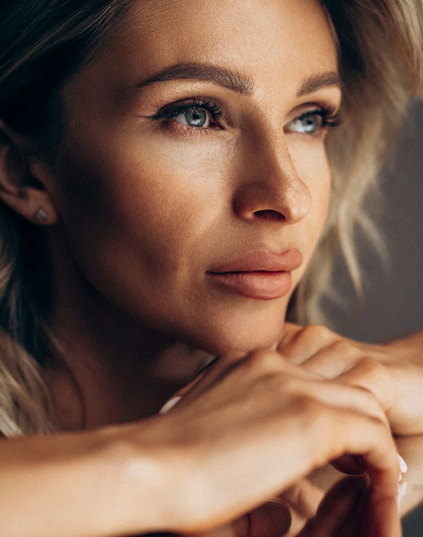 Close-up of a blonde woman with loose waves framing her face. Her skin is smooth with a light tan tone, and her expression is contemplative as she gazes slightly right. She has light grey-blue eyes, well-defined brows, and soft pink-nude lips. Subtle highlights accentuate her cheekbones and forehead. Small stud earrings are visible, and her hands rest gently on her chest. The muted gray-brown background and soft lighting emphasize her features. - RF Microneedling in Los Angeles, CA