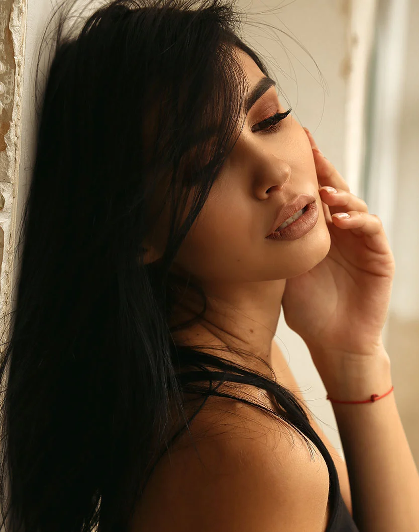 Close-up of a woman with dark hair framing her face and a strand across her forehead. Her eyes are closed, makeup soft and natural with rosy lips. One hand rests gently below her cheek, showing a thin red bracelet. She leans against a light-colored wall with a softly blurred background and warm, diffused lighting. - Sciton Profactional Laser in Los Angeles, CA