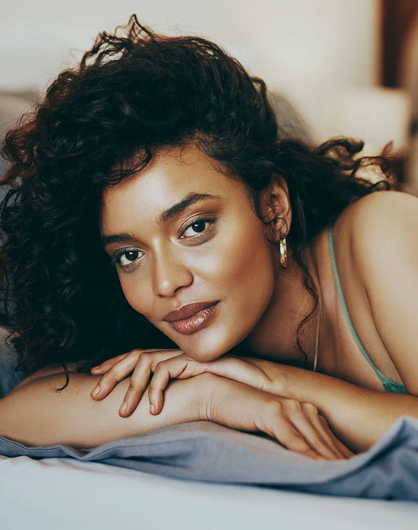 A woman with dark curly hair lies on a bed, looking calmly at the camera. She wears a light teal tank top, with her hands resting gently beside her face. Her light brown skin contrasts softly with the muted gray-blue bedding under warm, gentle lighting. - Soft Threads in Los Angeles, CA