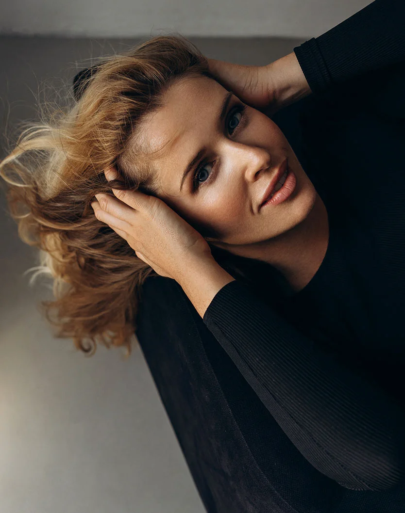 A woman with light brown, tousled hair is viewed from an overhead, slightly angled perspective. She holds her hair with both hands, her head tilted back slightly, and her expression relaxed. She has light skin and light blue eyes. She wears a fitted black top, and the background is a muted gray surface with a soft-focus effect. - Temple Augmentation in Los Angeles, CA